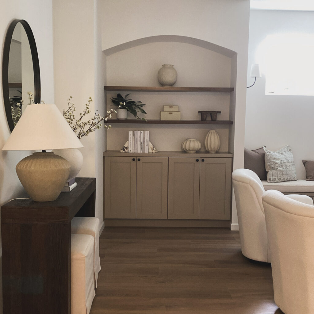 An image of a interior room with an arched built-in shelf with cabinetry, chairs, a window bench, a round mirror, a wood console table, lamp, and miscellaneous decorations.